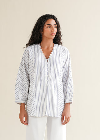 Striped Kimono Blouse – NW-05-A for Women – Summer Reverie Collection | KOA | Shop Online.
A woman modeling a striped kimono blouse with a V-neck, center front placket with buttons, and oversized kimono sleeves. The blouse is lilac with a pattern of darker stripes, and she is wearing white pants.