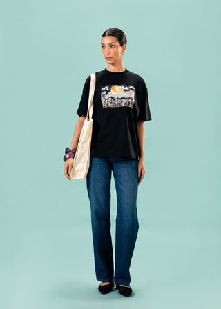 T-Shirt – Moon over Metropolis by Minhas Alee – WS25-215 for Women – Art Board 01 Collection | KOA | Shop Online.
Woman wearing a black t-shirt with a graphic design and blue jeans, standing against a light blue background.