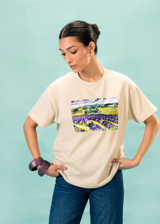 T-Shirt – The Purples Within by Ariba Choudhary – WS25-216 for Women – Art Board 01 Collection | KOA | Shop Online.
Woman wearing a beige t-shirt with a colorful graphic on a light blue background