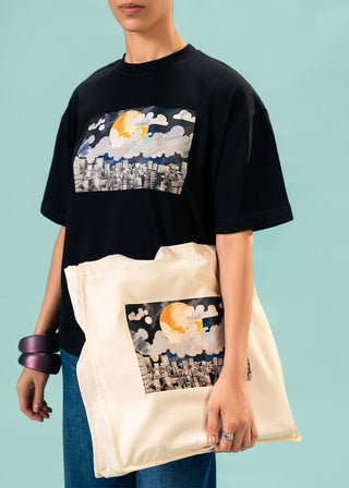 Tote Bag – Moon Over Metropolis by Minhas Alee – WS25-240 for Women – Art Board 01 Collection | KOA | Shop Online.
Women wearing a black t-shirt with a graphic design, holding a tote bag matching the design.
