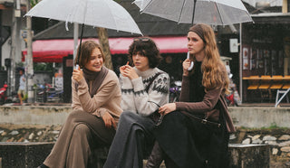 Three women sitting together under umbrellas on a street - wear KOA winter coded outfits.