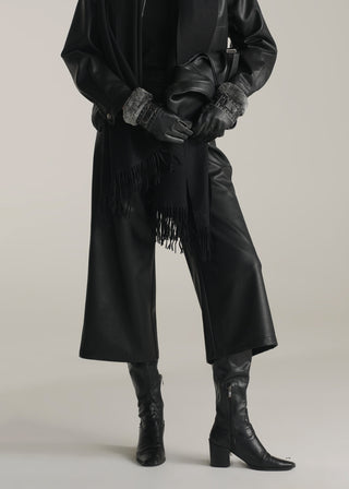 Women wearing a long black coat with fringes, black gloves, and black KOA leather pants on a plain background